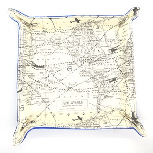 May include: A square fabric tray with a world map design in cream and black, featuring airplanes. The tray has a blue border and is labeled "THE WORLD" with Mercator Projection details. A decorative and functional item.