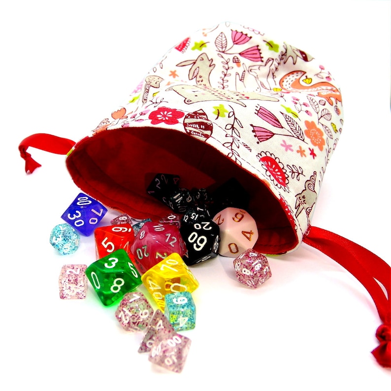 May include: A fabric dice bag with a red interior and a white exterior patterned with pink flowers, rabbits, and other floral elements. A variety of colorful dice are spilling out of the bag, including blue, red, green, and clear dice.