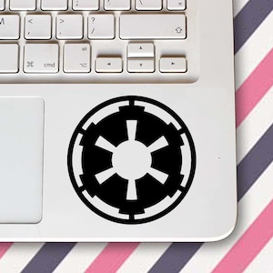 Könnte beinhalten: Ein schwarz-weißer Imperial-Symbol-Aufkleber befindet sich auf einem weißen Laptop. Der Laptop steht auf einer Oberfläche mit einem rosa, weißen und lila Streifenmuster.