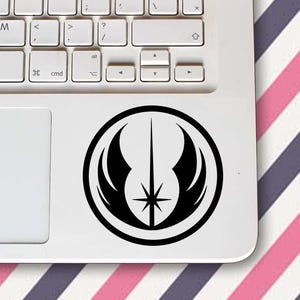 Pode incluir: Um autocolante de vinil a preto e branco com um design circular, uma estrela central e asas estilizadas. O autocolante está afixado num computador portátil branco. O teclado é visível. O fundo tem riscas diagonais cor-de-rosa e roxas.