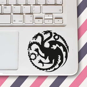 Puede incluir: Una calcomanía negra con un emblema de dragón de tres cabezas en una computadora portátil blanca. La computadora portátil está sobre una superficie con un patrón de rayas rosas, blancas y moradas.