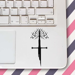 May include: A black vinyl decal featuring a tree and sword design is on a white laptop. The tree has many branches and is above the sword. The laptop is on a surface with pink, white, and purple diagonal stripes.