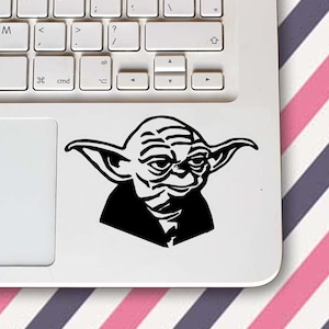 May include: A black and white Yoda sticker on a white laptop. The sticker depicts Yoda's face, with his distinctive ears and facial features. The laptop's keyboard is visible. The laptop is on a surface with diagonal stripes.