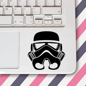 Può includere: Un adesivo in vinile in bianco e nero di un casco da Stormtrooper è attaccato a un laptop bianco. L'adesivo si trova in basso a destra del laptop, vicino al touchpad. Lo sfondo è un motivo a strisce diagonali rosa, bianche e grigie.