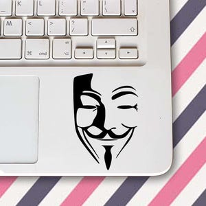 Könnte beinhalten: Ein schwarz-weißer Aufkleber der Guy-Fawkes-Maske, ein Symbol des Protests, befindet sich auf einem weißen Laptop. Die Tastatur des Laptops ist sichtbar. Der Hintergrund ist rosa, weiß und lila gestreift.