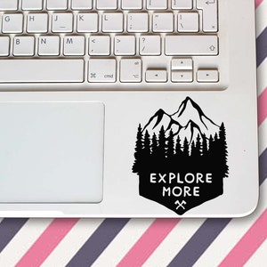 Puede incluir: Adhesivo negro con una silueta de montaña y bosque sobre un portátil blanco. El adhesivo tiene las palabras "EXPLORE MORE" en blanco, con hachas cruzadas debajo. Se ven el teclado y el panel táctil del portátil.