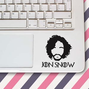 Könnte beinhalten: Ein schwarzer Vinyl-Aufkleber mit einer stilisierten Silhouette von Jon Snows Gesicht und dem Text "JON SNOW" darunter. Der Aufkleber befindet sich auf einem weißen Laptop, mit einer Tastatur darüber. Der Hintergrund ist ein rosa-weiß gestreiftes Muster.