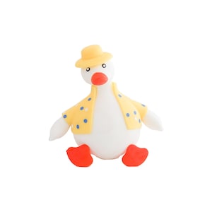 Puede incluir: Una caprichosa figura de pato blanco con sombrero amarillo y camisa amarilla con lunares azules. El pato tiene pico y patas naranjas. La figura está sentada y tiene una expresión amigable.