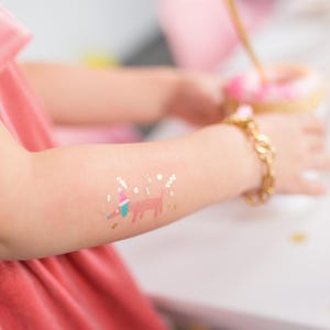May include: A child's arm with a temporary tattoo of a dog wearing a party hat. The tattoo is gold with pink, blue, and green accents. The tattoo is surrounded by gold glitter.
