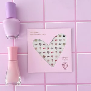 May include: Pink and purple nail polish bottles with a sheet of nail stickers featuring hearts, pretzels, rainbows, and the text "Love" in a heart shape. The sticker sheet is labeled "nail stickers" and "autocollants pour ongles". The sticker sheet also features the text "JORDAN SONDLER" and "DAYDREAM SOCIETY".