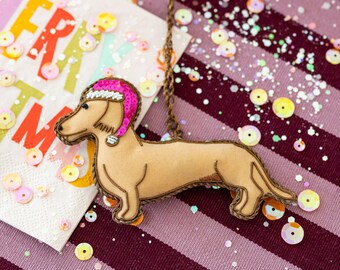 May include: A tan dachshund ornament wearing a pink Santa hat with a white trim. The dog is outlined in brown thread and hangs from a gold chain. The background includes a napkin with colorful text and sequins.