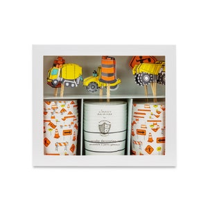 May include: Construction themed cupcake decorating set with orange and white cupcake liners, and yellow and orange construction vehicle toppers. The box includes the text "cupcake decorating set" and "ensemble à petits gâteaux".