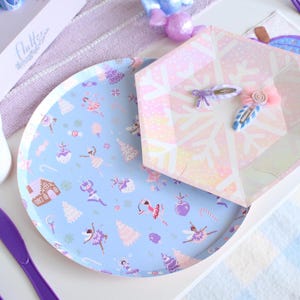 May include: A light blue paper plate with a ballerina and holiday-themed pattern, next to a snowflake-patterned box and two decorative hair clips. The plate features illustrations of ballerinas, gingerbread houses, and candy canes.