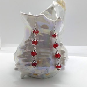 May include: A pair of long silver earrings with red glass beads. The earrings are hanging from a white ceramic seashell.