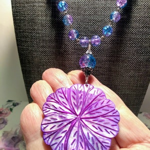 Pu&ograve; includere: Una collana con un ciondolo a forma di fiore di conchiglia madreperla viola, una catena d'argento e perline di vetro blu e viola.