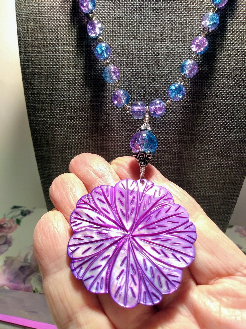 Pu&ograve; includere: Una collana con un ciondolo a forma di fiore di conchiglia madreperla viola, una catena d'argento e perline di vetro blu e viola.