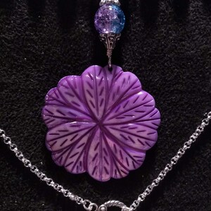 Pu&ograve; includere: Una collana con un ciondolo a forma di fiore viola, una catena d'argento e una chiusura. Il ciondolo &egrave; realizzato in conchiglia scolpita e presenta un design dettagliato. La catena &egrave; d'argento e ha una chiusura a scatto.