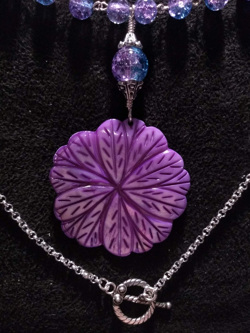 Pu&ograve; includere: Una collana con un ciondolo a forma di fiore viola, una catena d'argento e una chiusura. Il ciondolo &egrave; realizzato in conchiglia scolpita e presenta un design dettagliato. La catena &egrave; d'argento e ha una chiusura a scatto.