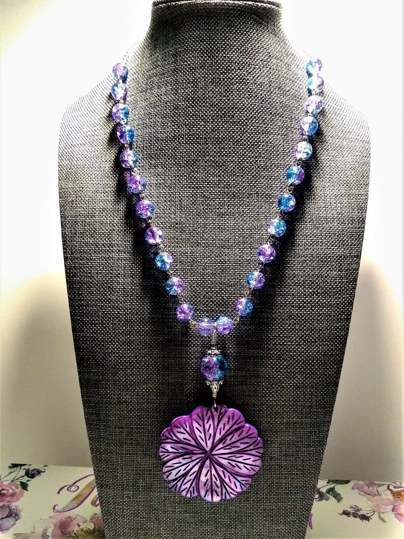 Pu&ograve; includere: Una collana di perline di vetro blu e viola con un grande ciondolo a forma di fiore viola. La collana &egrave; su un manichino grigio.