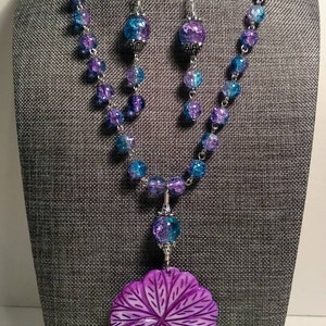 Pu&ograve; includere: Una collana di perline di vetro viola e blu con un grande ciondolo a forma di fiore viola. La collana &egrave; su un manichino e ha orecchini abbinati con un accento di perla.