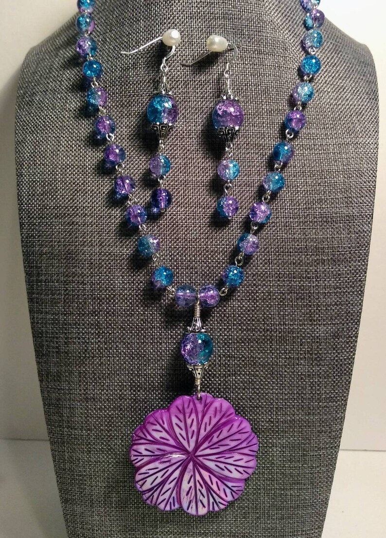 Pu&ograve; includere: Una collana di perline di vetro viola e blu con un grande ciondolo a forma di fiore viola. La collana &egrave; su un manichino e ha orecchini abbinati con un accento di perla.