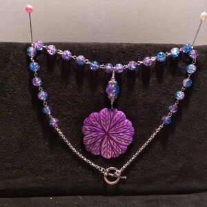 Pu&ograve; includere: Una collana con un ciondolo a forma di fiore viola, una catena d'argento e perline di vetro blu e viola.