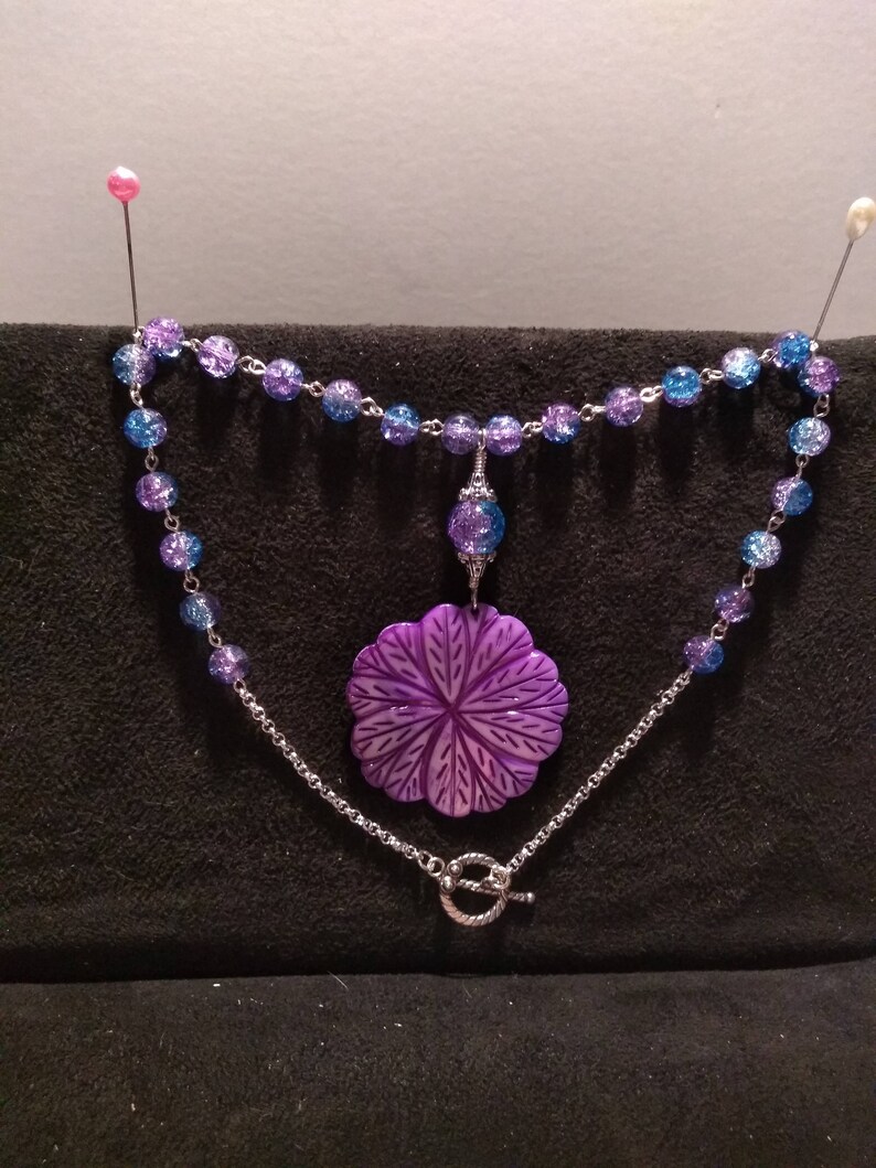 Pu&ograve; includere: Una collana con un ciondolo a forma di fiore viola, una catena d'argento e perline di vetro blu e viola.