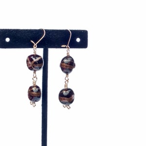 Puede incluir: Un par de pendientes colgantes con dos cuentas de vidrio negro y dorado en cada pendiente. Los pendientes están colgados de un expositor negro.