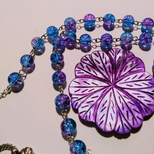Pu&ograve; includere: Una collana con un ciondolo a forma di fiore viola, una catena d'argento e perline di vetro blu e viola.