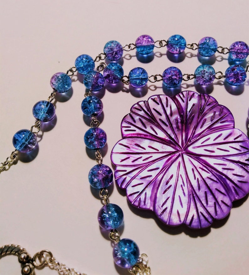 Pu&ograve; includere: Una collana con un ciondolo a forma di fiore viola, una catena d'argento e perline di vetro blu e viola.