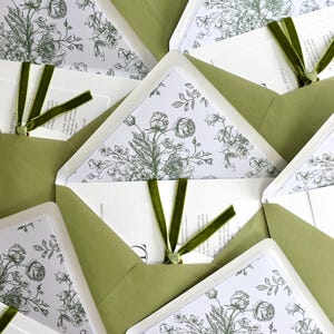 May include: An arrangement of olive green envelopes, each holding a floral invitation. The invitations display a botanical design in green tones and are tied with a dark green velvet ribbon. The envelopes are neatly arranged, creating an appealing visual pattern.