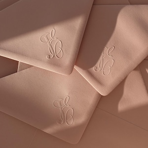 Puede incluir: Primer plano de varios sobres de color rosa claro con un diseño de monograma. Los sobres están dispuestos en un patrón en capas, con el monograma visible en las esquinas. La iluminación crea sombras y resalta la textura del papel.