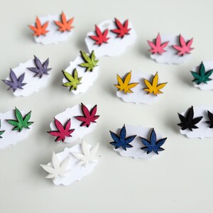 Puede incluir: Una colección de pendientes de botón con forma de hoja en varios colores, presentados en pequeñas tarjetas blancas. Los pendientes son de color naranja, rojo, rosa, morado, verde, amarillo, verde oscuro, azul, negro y blanco. Probablemente hechos de madera.