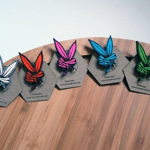May include: A collection of colorful enamel pins shaped like a stylized bunny head with marijuana leaves for ears. The pins are white, red, pink, blue, and green, each mounted on a gray hexagonal card. The website address www.joosings.com is printed on each card.