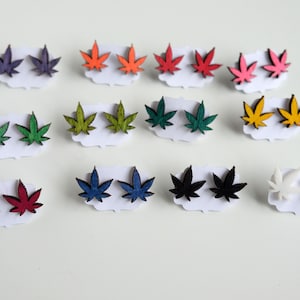 Puede incluir: Una colección de pendientes de botón con forma de hoja en varios colores. Los pendientes están dispuestos en pares e incluyen morado, naranja, rojo, rosa, verde, amarillo, azul, negro y blanco. Cada par se presenta en una pequeña tarjeta blanca.