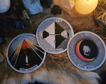 May include: Three circular cards with artistic illustrations. One shows a candle flame with rays, labeled THE FLAME and LI. Another features a black and white rose in a diamond, labeled ALETHEIA and LXXXVII. The third shows hands, labeled THE KISS and LX.