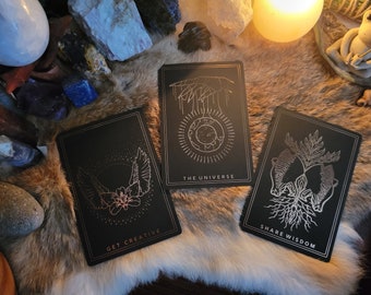 May include: Three rectangular black cards with gold foil line art illustrations. The left card shows wings and a flower with the text GET CREATIVE. The center card depicts a hand over a globe with THE UNIVERSE. The right card shows roots with SHARE WISDOM.