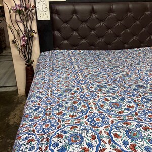 May include: A white cotton quilt with a blue and brown floral pattern. The quilt is spread out on a bed with a brown leather headboard.