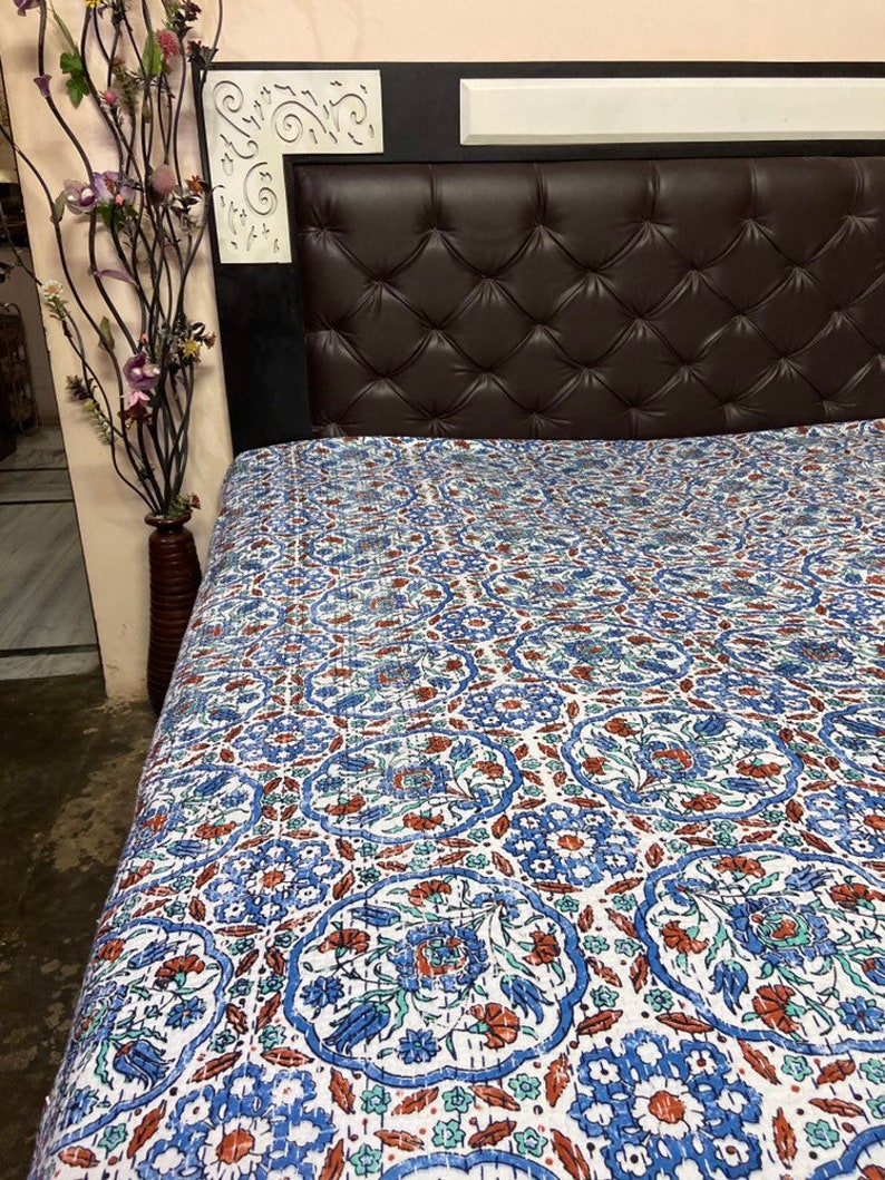 May include: A white cotton quilt with a blue and brown floral pattern. The quilt is spread out on a bed with a brown leather headboard.