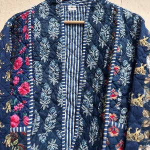 Peut inclure: Veste matelass&eacute;e avec un motif patchwork. La veste pr&eacute;sente un m&eacute;lange de motifs floraux, ray&eacute;s et imprim&eacute;s tigr&eacute;s dans des tons de bleu, de rose et de blanc. La veste est suspendue &agrave; un cintre en bois.