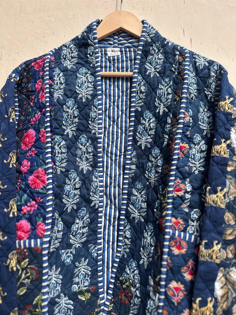 Peut inclure: Veste matelass&eacute;e avec un motif patchwork. La veste pr&eacute;sente un m&eacute;lange de motifs floraux, ray&eacute;s et imprim&eacute;s tigr&eacute;s dans des tons de bleu, de rose et de blanc. La veste est suspendue &agrave; un cintre en bois.