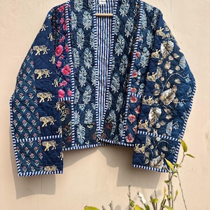 May include: A quilted jacket in shades of navy blue, featuring a patchwork design with floral, tiger, and leaf patterns. The jacket has a striped center panel and white trim. It is hanging on a wooden hanger.