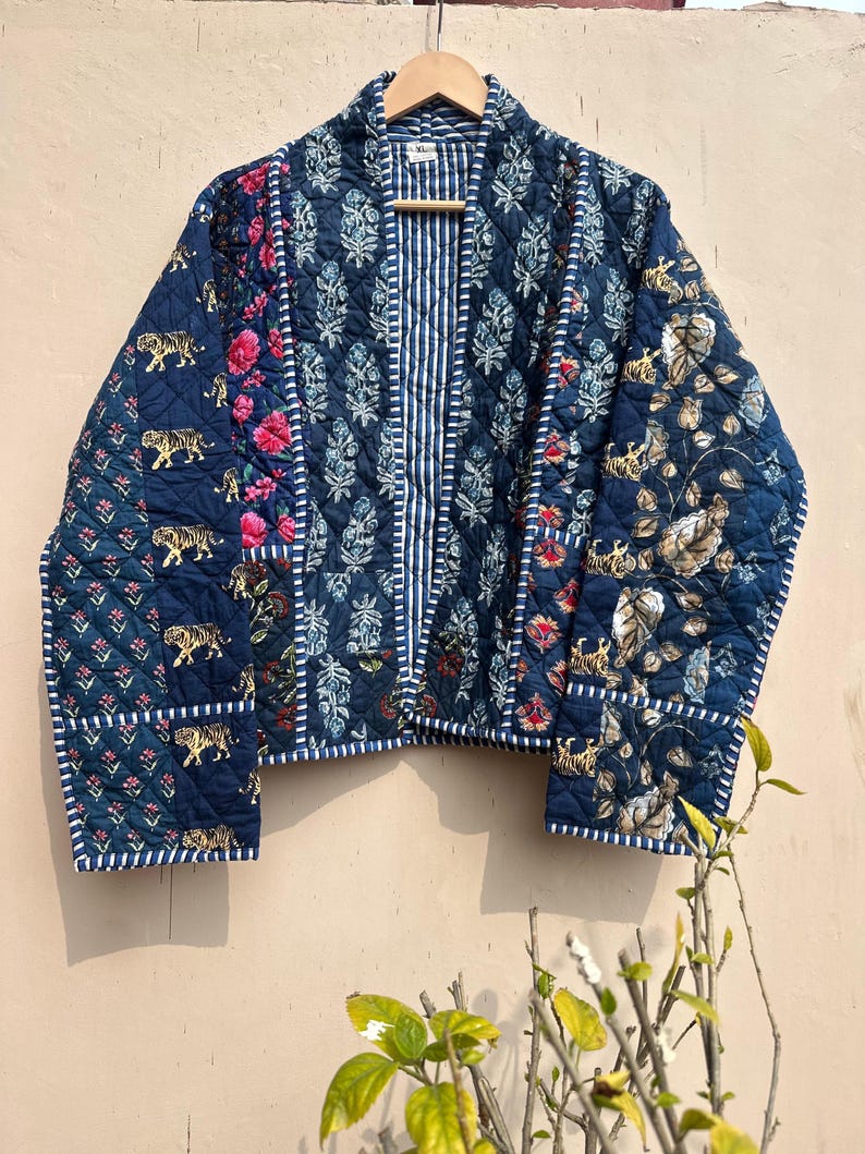 Peut inclure: Veste matelass&eacute;e bleu marine avec un motif patchwork. Elle pr&eacute;sente des motifs floraux, de tigres et de feuilles, un panneau central ray&eacute; et une bordure blanche. Suspendue &agrave; un cintre en bois.
