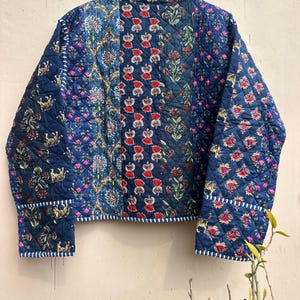 Peut inclure: Veste matelass&eacute;e bleue avec un motif patchwork. La veste pr&eacute;sente des motifs floraux et animaliers rouges et bleus. Elle a une texture matelass&eacute;e et est bord&eacute;e d'un tissu ray&eacute; blanc et bleu. Les manches et le col sont &eacute;galement orn&eacute;s de la m&ecirc;me garniture ray&eacute;e.