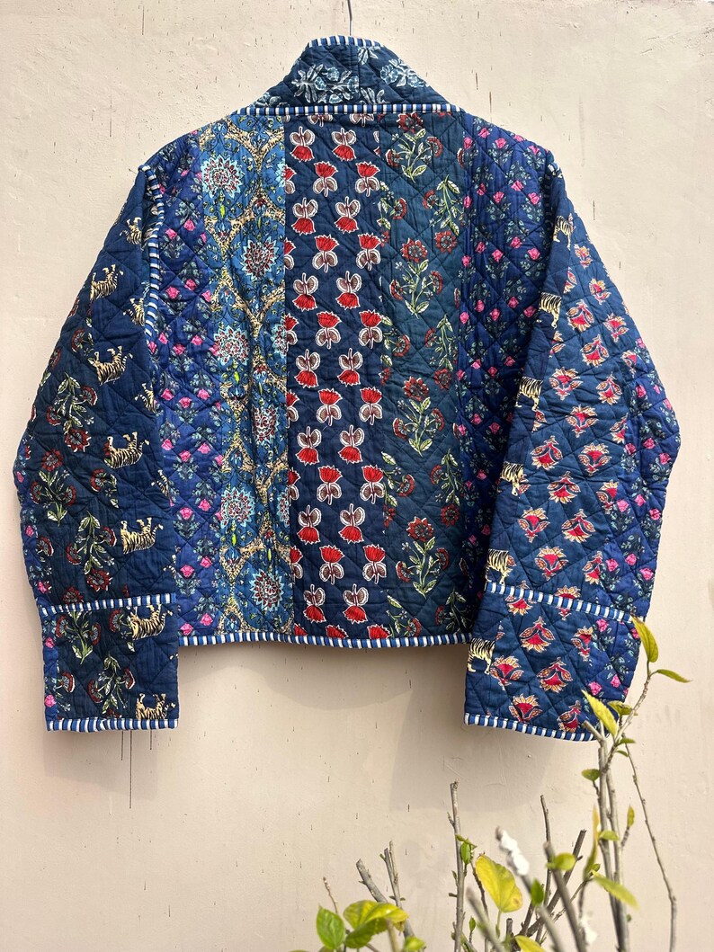 Peut inclure: Veste matelass&eacute;e bleue avec un motif patchwork. La veste pr&eacute;sente des motifs floraux et animaliers rouges et bleus. Elle a une texture matelass&eacute;e et est bord&eacute;e d'un tissu ray&eacute; blanc et bleu. Les manches et le col sont &eacute;galement orn&eacute;s de la m&ecirc;me garniture ray&eacute;e.