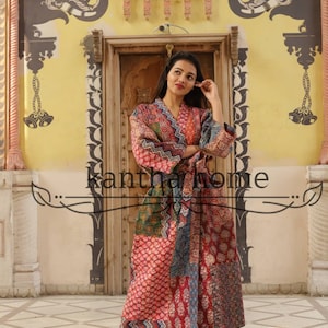 Könnte beinhalten: Eine Frau trägt eine lange, bunte Patchwork-Robe mit Blumenmuster. Die Robe ist in der Taille mit einem Band gebunden. Die Frau steht vor einer Tür mit einem dekorativen Torbogen. Der Text "Kantha Home" ist im Bild sichtbar.