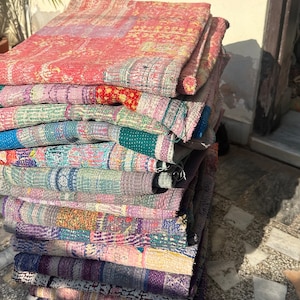 Könnte beinhalten: Ein Stapel bunter Kantha-Steppdecken, hergestellt aus recycelten Saris, mit aufwendigen Patchwork-Mustern. Die Decken sind gefaltet und auf einer Steinoberfläche gestapelt.
