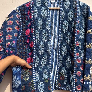Peut inclure: Veste matelass&eacute;e bleue orn&eacute;e de motifs floraux et ray&eacute;s. La veste a une coupe d&eacute;contract&eacute;e, avec un m&eacute;lange de panneaux floraux et ray&eacute;s. La veste est suspendue &agrave; un cintre en bois.