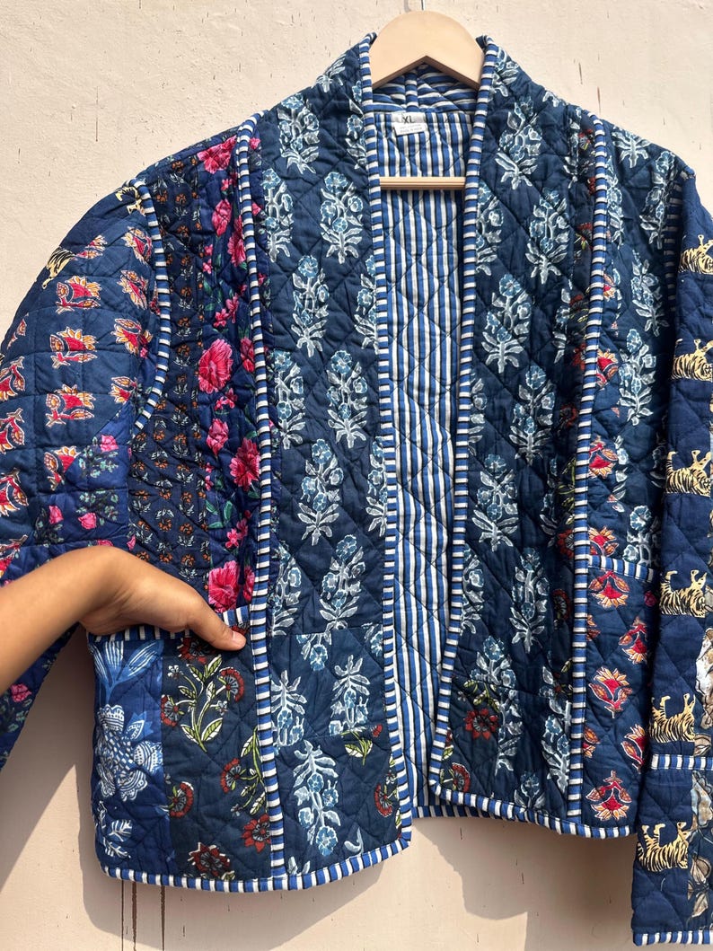 Peut inclure: Veste matelass&eacute;e bleue orn&eacute;e de motifs floraux et ray&eacute;s. La veste a une coupe d&eacute;contract&eacute;e, avec un m&eacute;lange de panneaux floraux et ray&eacute;s. La veste est suspendue &agrave; un cintre en bois.