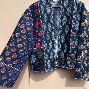 Peut inclure: Veste matelass&eacute;e bleu marine avec patchwork &agrave; imprim&eacute; floral et tigre. La veste pr&eacute;sente un panneau central ray&eacute; et une bordure blanche. Les manches sont longues et la veste est suspendue &agrave; un cintre en bois.